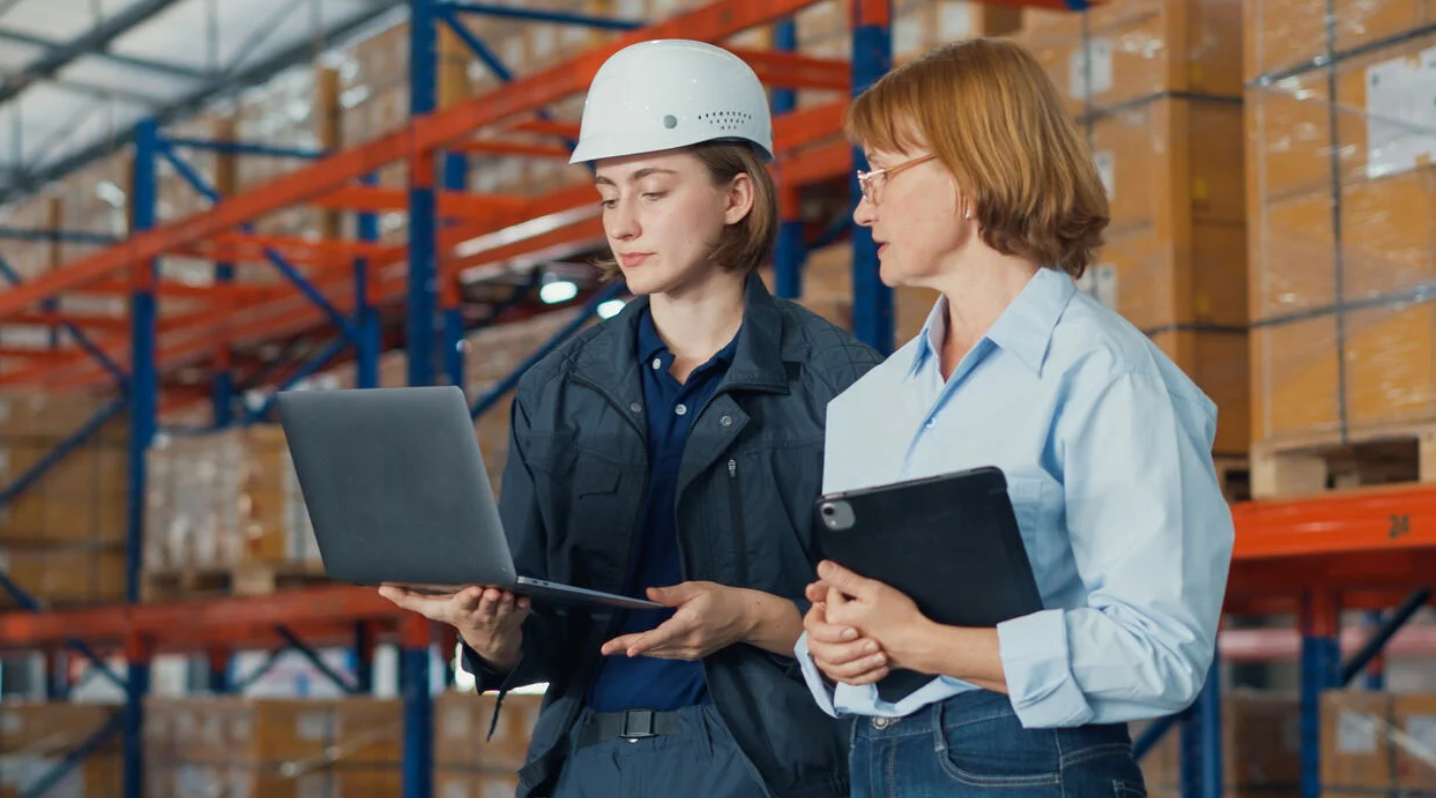 Two engineers in a factory setting, reviewing data on a laptop while collaborating on a project or process.