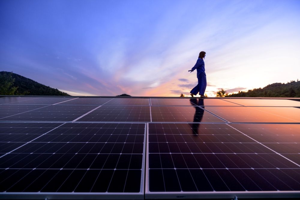 Person in blue overalls walking on a rooftop covered with solar panels at sunset, with hills and a colorful sky in the background