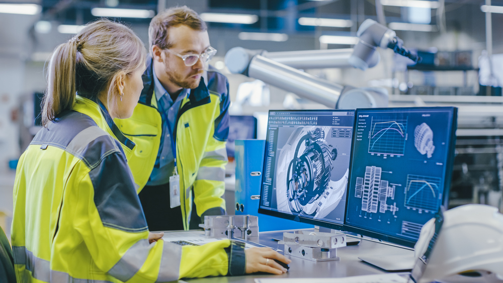 Two workers analyzing data on a laptop in a high-tech manufacturing facility, surrounded by automated machinery and industrial equipment.