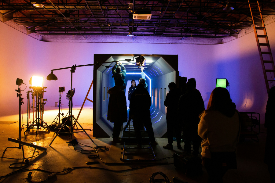 A film crew is gathered around a large screen displaying a digital tunnel. Lighting equipment and a ladder are visible in the studio setting.
